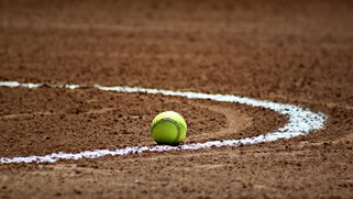 A yellow baseball on the clay of a baseball field.