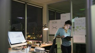 A woman working late at night in her office, holding a tablet in her hands.