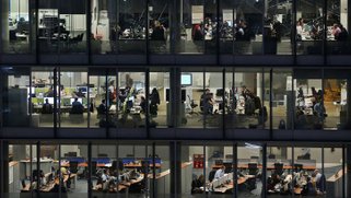 Picture of a building with big glass windows showing a crowded workspace at night.